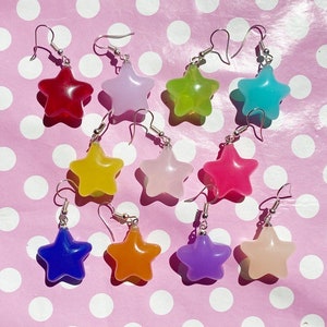 May include: A set of ten star-shaped earrings in various colours, including red, pink, yellow, green, blue, purple, and orange. The earrings are made of clear resin and have silver hooks.