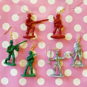 Puede incluir: Un par de pendientes con soldados de juguete de plástico en miniatura. Los pendientes están disponibles en tres colores: rojo, verde y plateado. Cada soldado sostiene un arma y está de pie sobre una pequeña base.