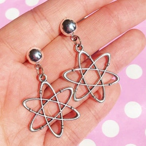 Może przedstawiać: Para srebrnych kolczyków w kształcie atomu z okrągłą srebrną koralikiem na górze każdego kolczyka.