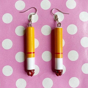 Op de afbeelding: Een paar novelty oorbellen in de vorm van sigaretten. Elke oorbel heeft een gele cilinder met een rode band, een wit gedeelte en een rood, getextureerd uiteinde, opgehangen aan zilverkleurige oorhaakjes.