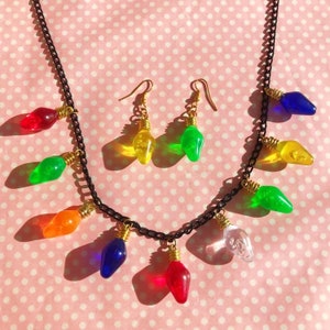 May include: A necklace and earrings set featuring colorful light bulb charms. The necklace has a black chain with multiple light bulb charms in various colors, including red, green, and blue. The earrings have a gold hook and a single light bulb charm.