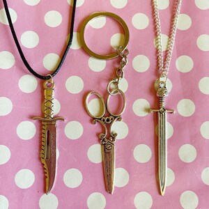 Może przedstawiać: Trzy metalowe wisiorki na różowym tle w kropki. Wisiorek w kształcie sztyletu na czarnym sznurku, brelok do kluczy w kształcie nożyczek i wisiorek w kształcie miecza na srebrnym łańcuszku. Sztylet ma tekst na ostrzu.