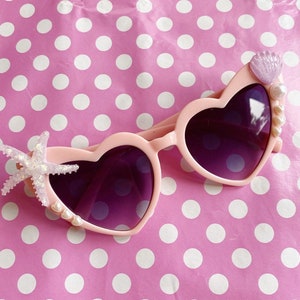 May include: Pink heart-shaped sunglasses with purple lenses, decorated with a starfish, seashells, and pearls.