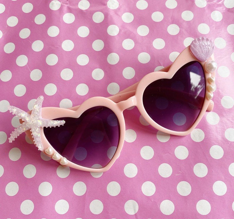 May include: Pink heart-shaped sunglasses with purple lenses, decorated with a starfish, seashells, and pearls.