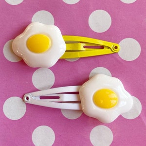 May include: Two hair clips with a fried egg design. Each clip has a white egg with a yellow yolk. One clip is on a yellow barrette, and the other is on a white barrette. The background is pink with white polka dots.