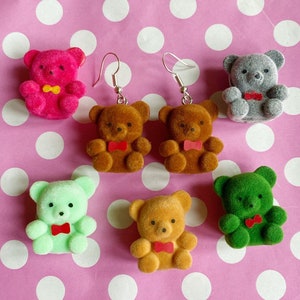 May include: Six pairs of dangle earrings featuring plush teddy bear charms in various colours, including pink, brown, grey, and green. Each bear charm has a small red bow tie.