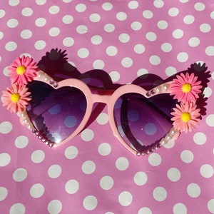 May include: Pink heart-shaped sunglasses with purple lenses and flower embellishments on the sides. The sunglasses are decorated with rhinestones and pearls.