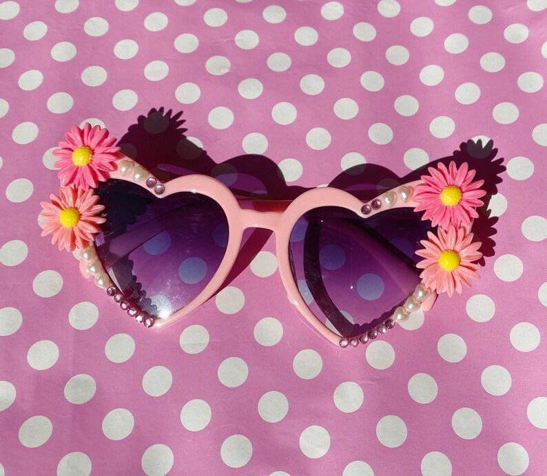 May include: Pink heart-shaped sunglasses with purple lenses and flower embellishments on the sides. The sunglasses are decorated with rhinestones and pearls.