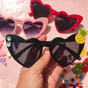 May include: Black heart-shaped sunglasses with rhinestones and a pineapple charm. The sunglasses have a black frame and dark lenses.