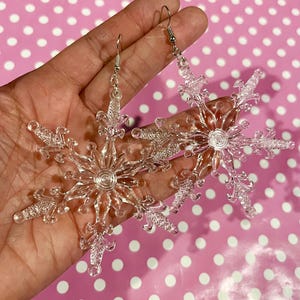 May include: A pair of clear, snowflake-shaped earrings with silver hooks. The earrings are made of a transparent material and feature detailed designs, resembling ice crystals. The earrings are held in a hand against a pink background with white polka dots.