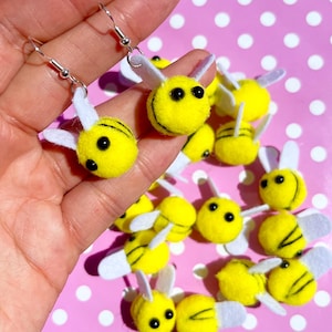Könnte beinhalten: Ein Paar Ohrhänger mit gelben Pompon-Bienen, die schwarze Streifen, weiße Filzflügel und schwarze Perlenaugen haben, gehalten vor einem rosa Hintergrund mit weißen Punkten, umgeben von weiteren losen Bienen-Anhängern.