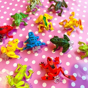 Puede incluir: Una colección de ranas de juguete coloridas en varios tonos de verde, amarillo, rojo y azul. Las ranas están dispuestas sobre un fondo rosa con lunares blancos. Cada rana está sujeta a un clip de plástico transparente.