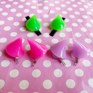 May include: Five pairs of plastic devil horns hair clips. Two pairs are green, two pairs are pink, and one pair is purple.