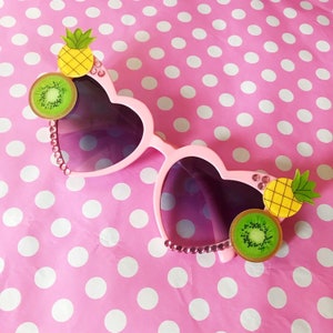 May include: Pink heart-shaped sunglasses with rhinestones and fruit accents. The sunglasses have a kiwi and pineapple on each side.