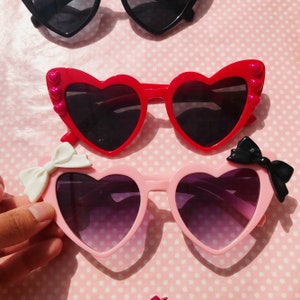 May include: Two pairs of heart-shaped sunglasses with bows. The top pair is red with pink hearts on the sides. The bottom pair is pink with a white bow on the left side and a black bow on the right side.