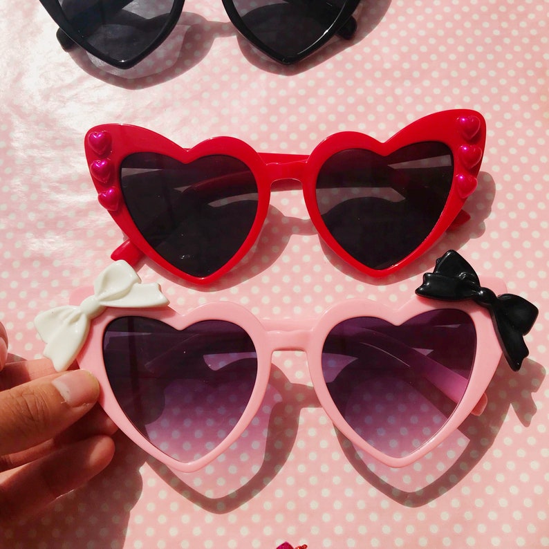 May include: Two pairs of heart-shaped sunglasses with bows. The top pair is red with pink hearts on the sides. The bottom pair is pink with a white bow on the left side and a black bow on the right side.