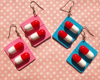 May include: A pair of earrings featuring a red and white capsule design. The earrings are made of wood and have a silver hook. The background is a pink polka dot pattern.