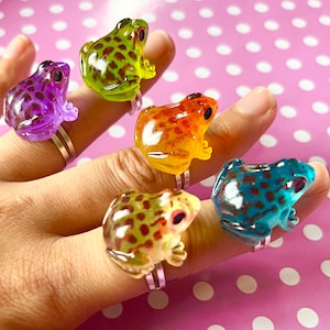 Op de afbeelding: Een verzameling kleurrijke kikkerringen in paars, groen, oranje, geel en blauw. Elke ring heeft een gedetailleerd kikkerontwerp met een zilveren band, perfect om een ​​vleugje fantasie toe te voegen.