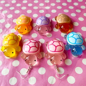 May include: Set of ten colourful turtle earrings. Each earring features a different coloured turtle with a detailed shell design. The turtles are made of a translucent material and have silver earring hooks.