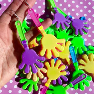May include: Novelty earrings with a hand-shaped design in vibrant colours. The earrings are yellow, purple, and green with pink and green handles. The earrings are made of plastic and have silver-coloured hooks.