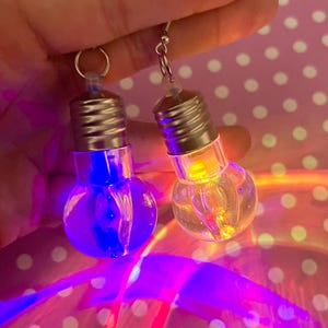 May include: A pair of dangling earrings shaped like miniature light bulbs. One bulb glows with a vibrant blue light, while the other emits a warm yellow light. Each earring features a silver-toned metal screw base and a clear glass bulb.