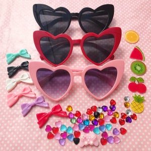 May include: Three pairs of heart-shaped sunglasses in black, red, and pink. The sunglasses are arranged in a row, with the black pair on top, the red pair in the middle, and the pink pair on the bottom. There are also various colorful decorations scattered around the sunglasses, including bows, fruit, and rhinestones.