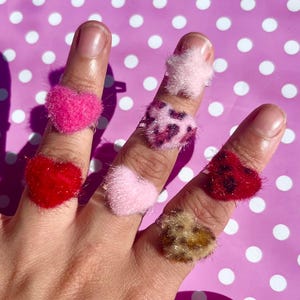 May include: A hand displaying several fuzzy heart-shaped rings in various colors, including pink, red, and leopard print. The rings are worn on different fingers, creating a playful and colorful display. The background is a pink surface with white polka dots.