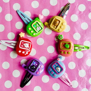May include: Six colourful hair clips with a retro handheld game design. Each clip features a different colour and a small, cartoon animal or object inside the game screen. The clips are arranged on a pink polka dot background.