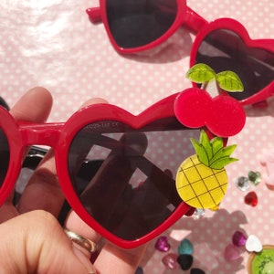 May include: Red heart-shaped sunglasses with black lenses and fruit-shaped embellishments. A cherry and pineapple are attached to the right side of the sunglasses.