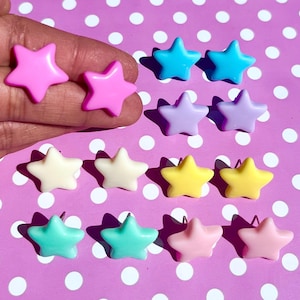 Könnte beinhalten: Eine Auswahl an sternförmigen Ohrsteckern in Pastellfarben wie Rosa, Blau, Gelb und Grün. Die Ohrringe liegen auf einem lila Hintergrund mit weißen Punkten.