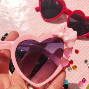 May include: Pink heart-shaped sunglasses with a pink bow and glitter heart accents. The sunglasses have dark lenses.