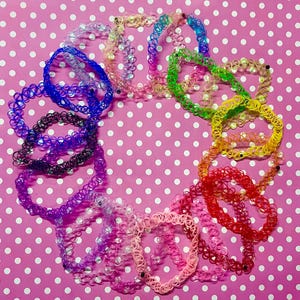 Puede incluir: Una colección de collares gargantilla elásticos y coloridos en varios tonos, incluyendo azul, verde, amarillo, rojo y rosa. Los collares están dispuestos en un patrón circular sobre un fondo rosa con lunares blancos. Accesorio retro.