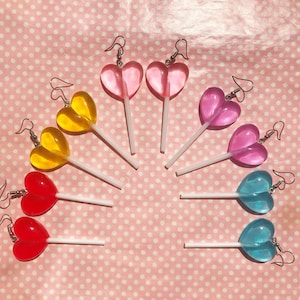 Könnte beinhalten: Ohrringe in Form von herzförmigen Lollis in Rot, Gelb, Rosa, Lila und Blau. Jeder Ohrring hat ein klares, glänzendes Herz, das an einem weißen Stiel und silberfarbenen Ohrhaken befestigt ist. Die Ohrringe sind auf einem rosa Hintergrund mit Polka Dots angeordnet.