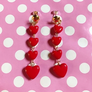 Könnte beinhalten: Ein Paar baumelnde Ohrringe mit roten herzförmigen Anhängern. Die Ohrringe haben einen goldfarbenen Stecker und eine Verbindungs-Kette. Der Hintergrund ist eine rosa Oberfläche mit weißen Punkten.