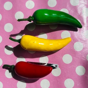 Könnte beinhalten: Drei Plastik-Chili-Haarklammern in Rot, Gelb und Grün. Die Klammern befinden sich auf einem rosa Hintergrund mit weißen Punkten.
