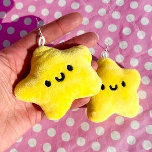 May include: A pair of yellow star-shaped plush earrings with a smiling face. The earrings are on a pink background with white polka dots.