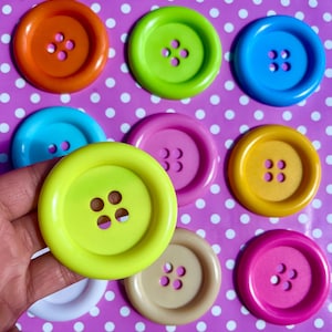 Puede incluir: Una colección de botones coloridos y de gran tamaño en tonos naranja, verde, azul, rosa, amarillo y beige. Cada botón tiene forma redondeada con cuatro o tres agujeros para coser. Los botones están dispuestos sobre un fondo morado con lunares blancos.