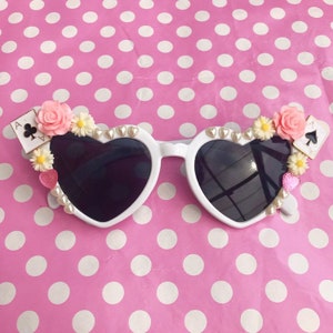 May include: White heart-shaped sunglasses with black lenses and decorative embellishments, including pink flowers, white flowers, pearl-like beads, and playing card charms. The sunglasses are on a pink and white polka dot background.