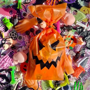 May include: A collection of Halloween-themed decorations, including an orange cellophane bag with a jack-o'-lantern face. The image features plastic hands, skeleton parts, and various spooky charms in orange, green, pink, and black.