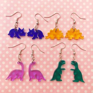 Könnte beinhalten: Ein Set mit sechs Paaren Ohrhängern mit verschiedenen Dinosauriersilhouetten. Die Ohrringe sind aus klarem Acryl und in Blau, Gelb, Grün und Lila erhältlich.