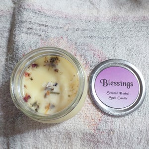 May include: A glass jar candle with a white wax base and dried herbs and flowers inside. The lid is silver with a purple label that reads "Blessings Scented Herbal Spell Candle".