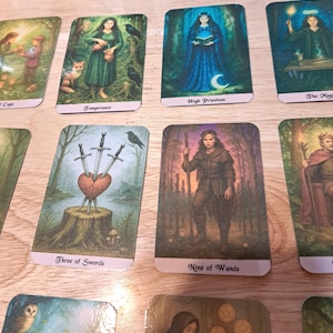 May include: A collection of tarot cards featuring various illustrations. The cards depict figures in natural settings, including a woman in a blue dress, a woman with a fox, and a heart pierced by swords. The cards are on a light wood surface.