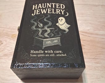 May include: A black wooden box with the words "HAUNTED JEWELRY" in white lettering. The box features a graphic of a treasure chest with smoke, a necklace, and a smiling ghost. The text "Handle with care. Some spirits are still.. attached." is also visible.
