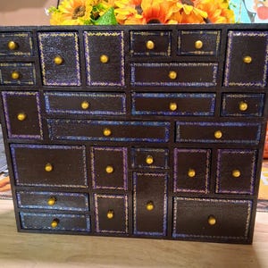 May include: A dark brown wooden storage chest featuring twenty-four drawers of varying sizes. Each drawer is accented with gold-colored knobs and decorative borders in shades of blue, purple, and gold, creating a detailed grid-like appearance.