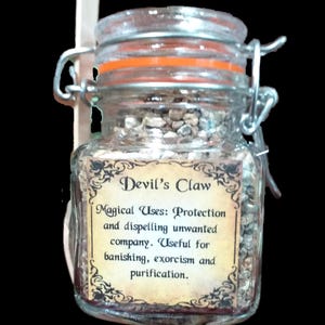 May include: A clear glass jar with a metal wire bail closure and an orange rubber seal, filled with small brown and grey botanical pieces. A label on the front reads: Devil's Claw, Magical Uses: Protection and dispelling unwanted company.