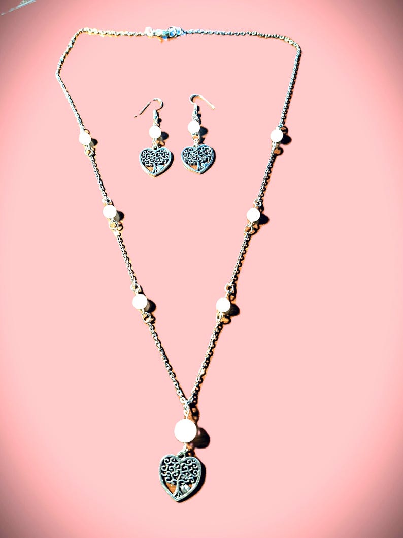 May include: A jewelry set consisting of a silver-toned chain necklace and matching drop earrings. The necklace features white beads interspersed along the chain, ending in a heart-shaped pendant with a tree design. The earrings mirror this heart motif.