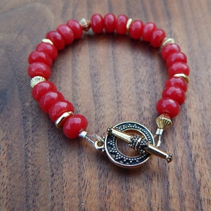 Könnte beinhalten: Ein rotes Perlenarmband mit goldenen Akzenten und einem dekorativen Verschluss. Das Armband hat runde, glänzende rote Perlen, die durch kleine goldfarbene Abstandshalter getrennt sind. Der Verschluss ist aus goldfarbenem Metall mit aufwendigen Details.