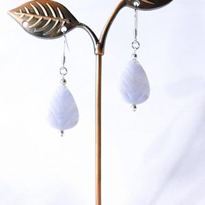 May include: A pair of light blue teardrop earrings with a leaf-like texture, hanging from silver hooks. The earrings are displayed on a bronze-colored jewelry stand with leaf-shaped accents.