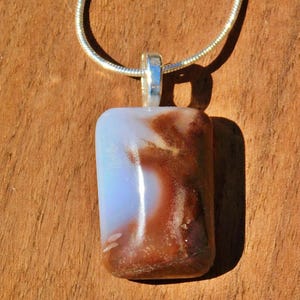 May include: A rectangular pendant with a silver bail hangs from a silver chain. The pendant features a blend of colors, including light blue, white, and brown. The pendant is approximately 1.5 inches long and 1 inch wide.