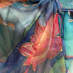 May include: Close-up of a sheer, flowing scarf with a hand-painted floral design. The scarf features a vibrant color palette of blues, pinks, and greens, with gold accents outlining the floral details. The fabric is knotted at the top.